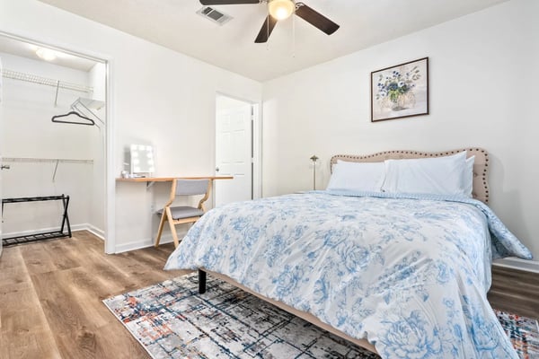 Bright bedroom with upholstered bed, blue floral bedding, wooden desk, ensuite bathroom, and ceiling fan